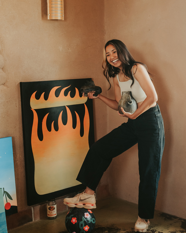 Artist and designer Paulina Ho stands next to one of her large-scale canvases, her arms full of ceramics.