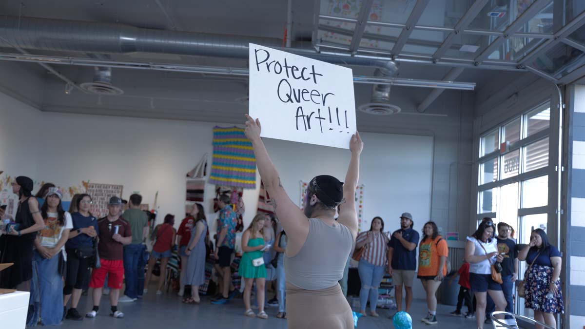 Performer holding a sign reading "Protect Queer Art!" at Lubbock's 5&J Gallery