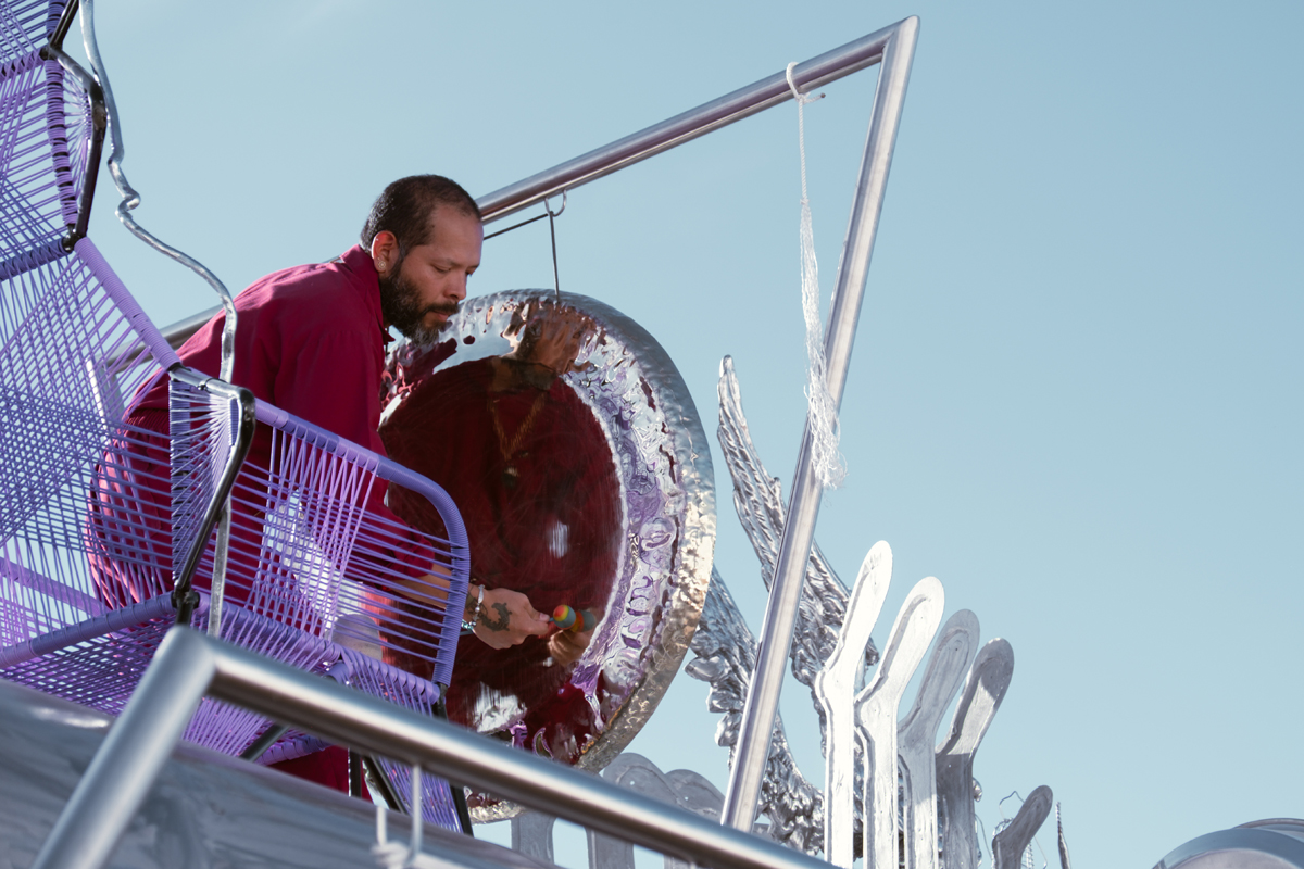 A man sits in a purple throne playing a gong. Guadalupe Maravilla Mariposa Relampago is a musical, winged bus with its own migration narrative.