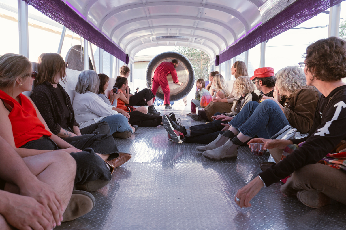 People line the sides of a bus, watching a man in a red jumpsuit play a gong. Guadalupe Maravilla Mariposa Relampago is a musical, winged bus with its own migration narrative.