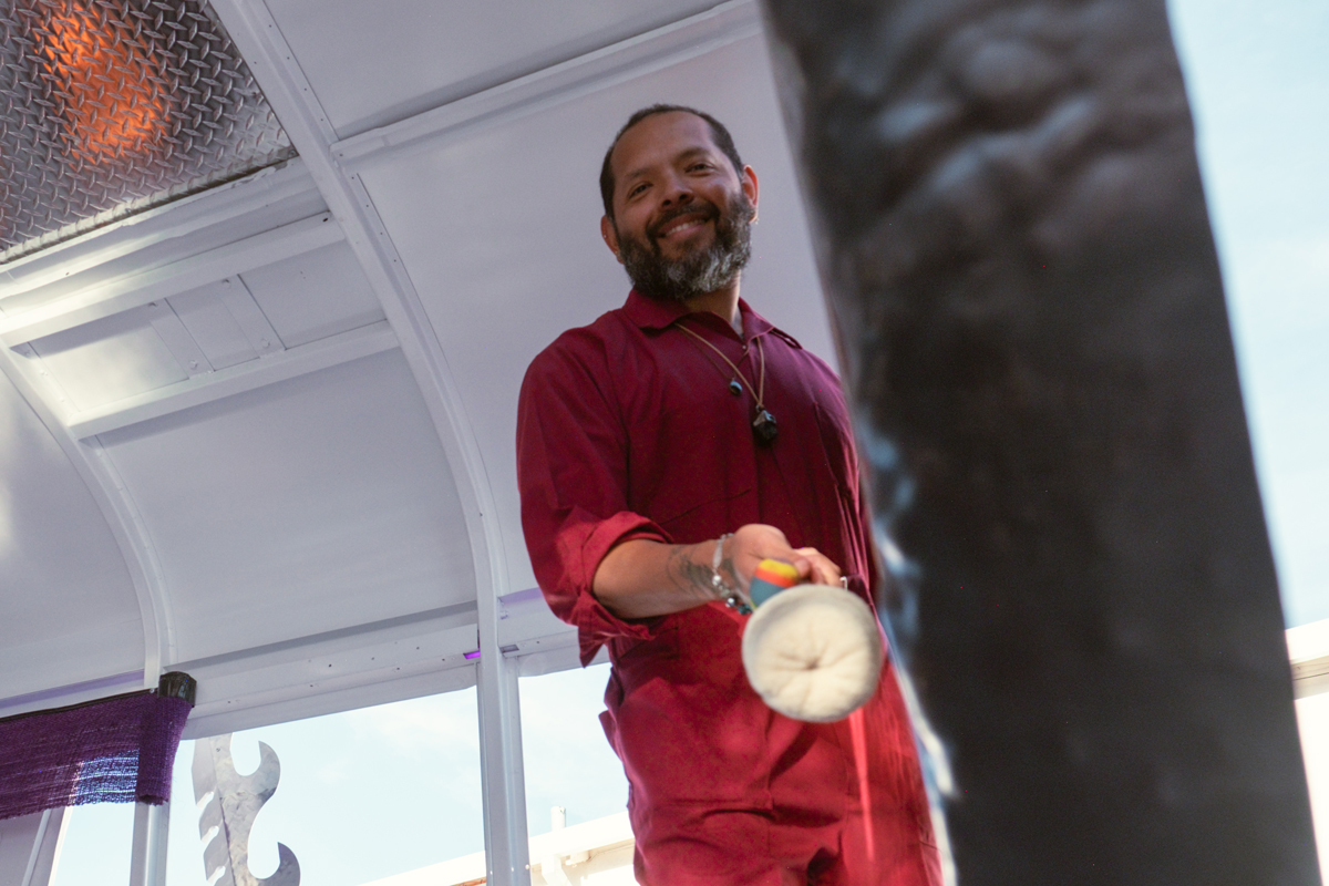 A man in a red jumpsuit smiles at the camera as he plays a gong inside a school bus. 