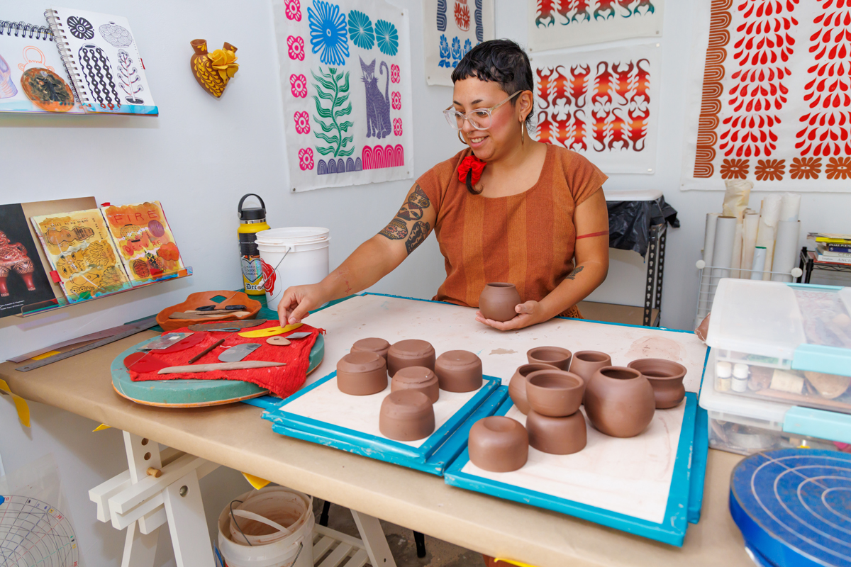 A ceramic artist sits in her studio, holding a ceramic vessel and selecting a tool. The Houston Center for Contemporary Craft is just one of the Southwest artist residencies featured in Southwest Contemporary's Fall 2024-Winter 2025 guide to forthcoming residency deadlines.