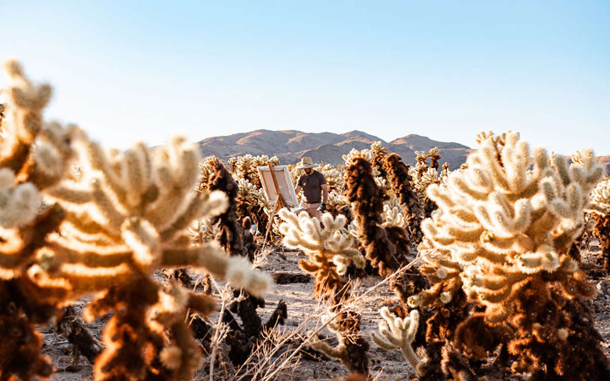 An artist paints among cholla cacti. The Joshua Tree Highlands Artist Residency is just one of the Southwest artist residencies featured in Southwest Contemporary's Fall 2024-Winter 2025 guide to forthcoming residency deadlines.