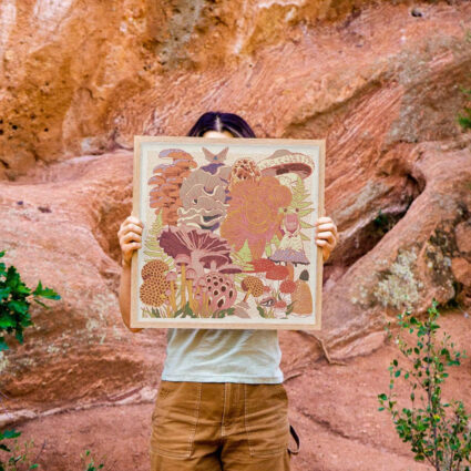 A woman holds a print with mushroom motifs in a red desert landscape.