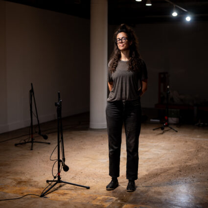 A woman stands in front of a patch of gritty earth on a concrete floor, with four microphones positioned around the room. Karima Walker is a Tucson-based artist with a major solo exhibition at MOCA Tucson.