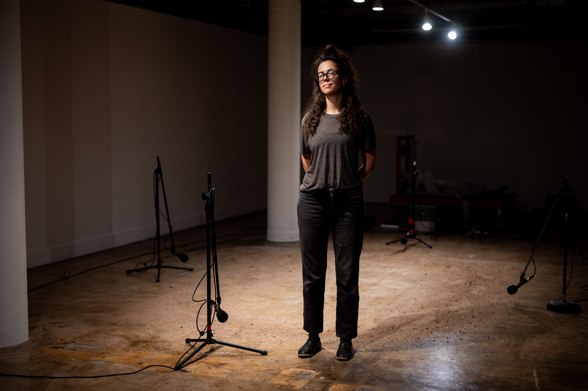 A woman stands in front of a patch of gritty earth on a concrete floor, with four microphones positioned around the room. Karima Walker is a Tucson-based artist with a major solo exhibition at MOCA Tucson.