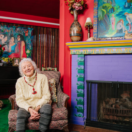 An artist sits among her paintings in her vibrant home.