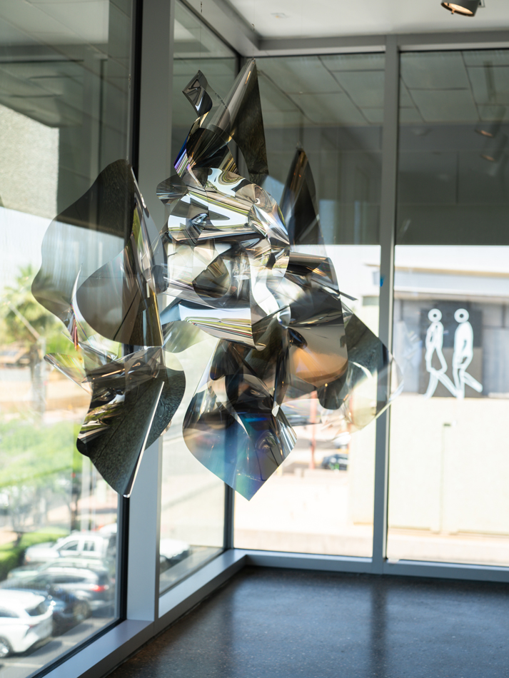 Installation view of a career retrospective by Larry Bell in Phoenix, featuring a suspended, squiggly, reflective silver sculpture.