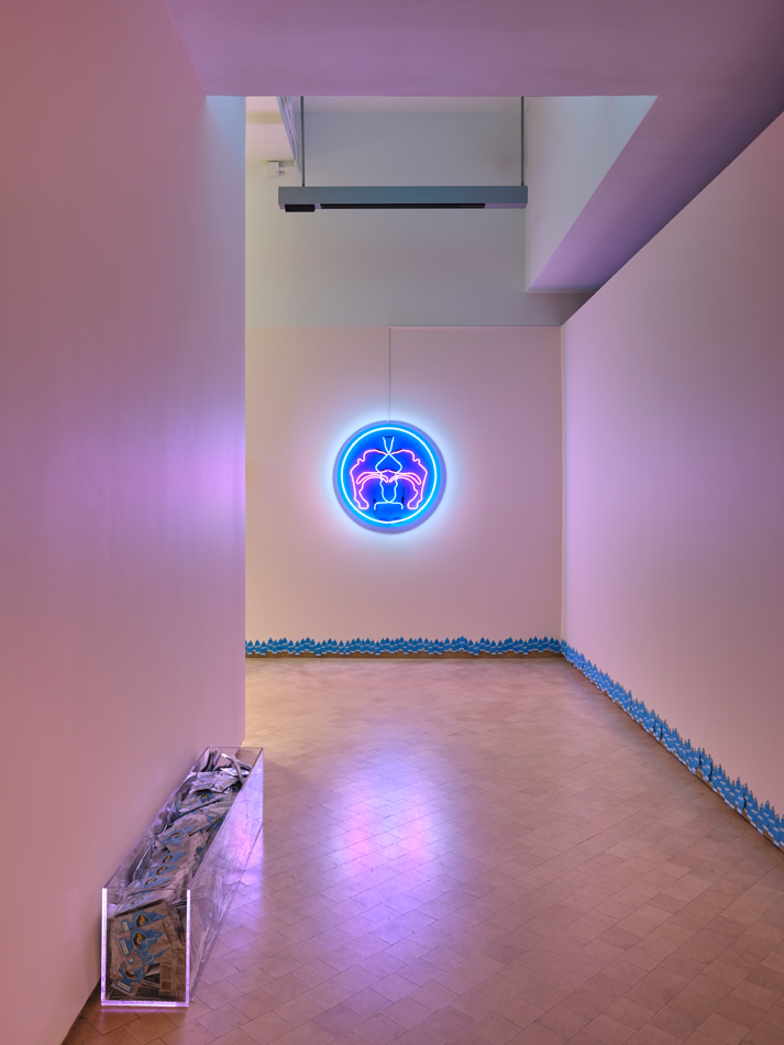 Installation view of a purple-and-blue neon sign glowing in a museum space. Curated by Maria Elena Ortiz.