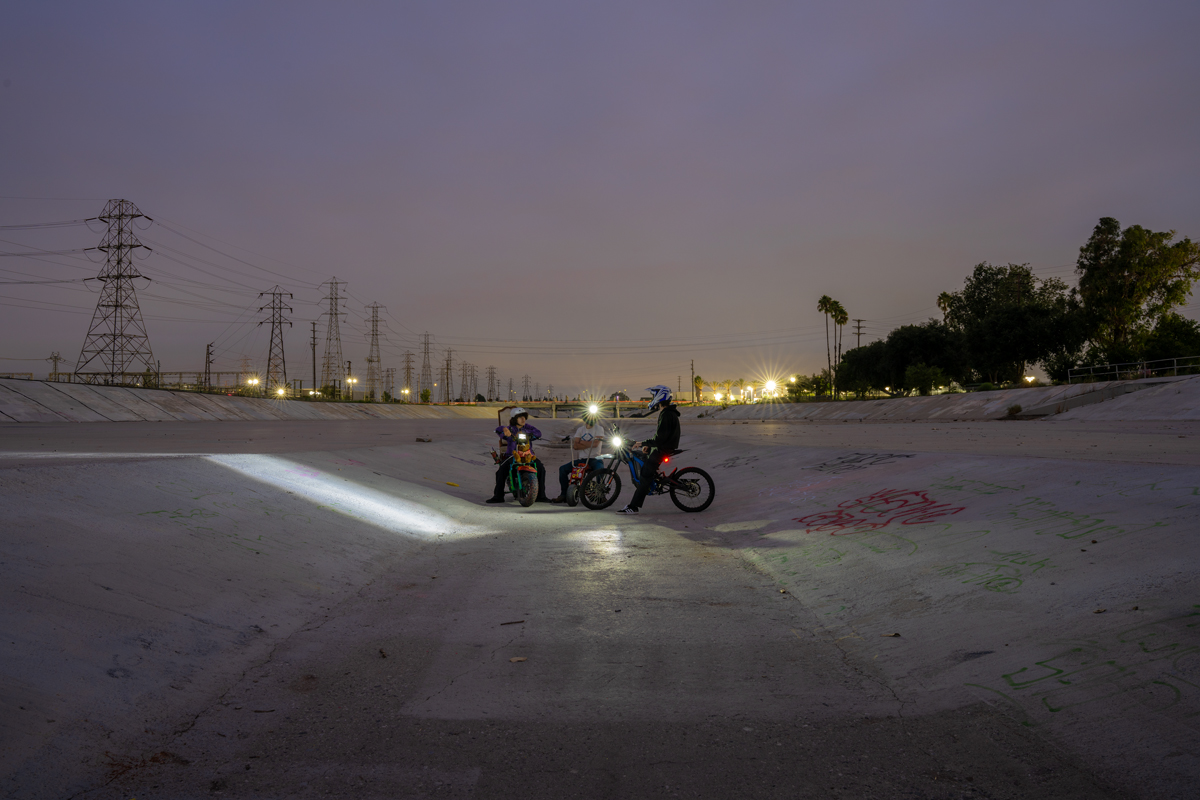 Night scene of people on bikes in a causeway, their flashlights lighting up the cement. Curated by Maria Elena Ortiz.