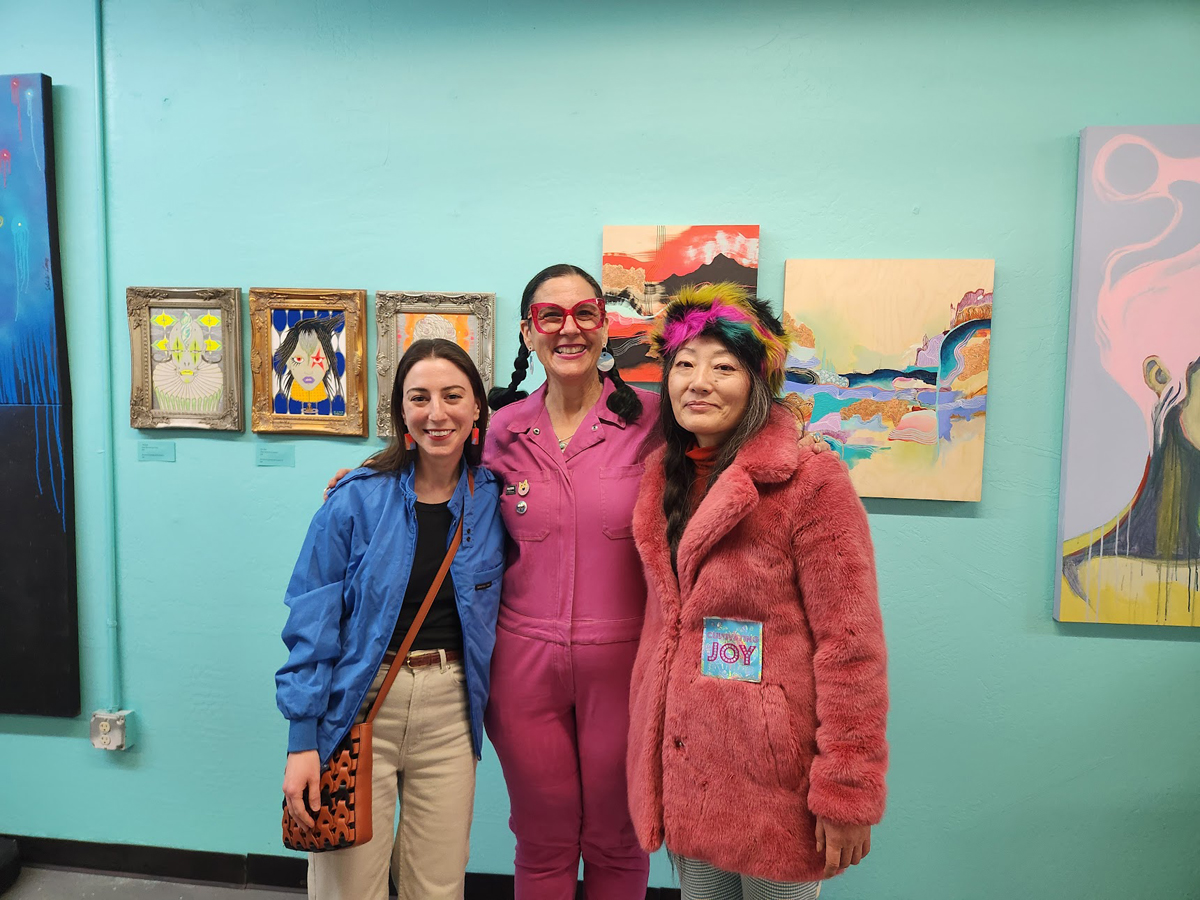 Three women pose in front of colorful artworks on a teal wall.