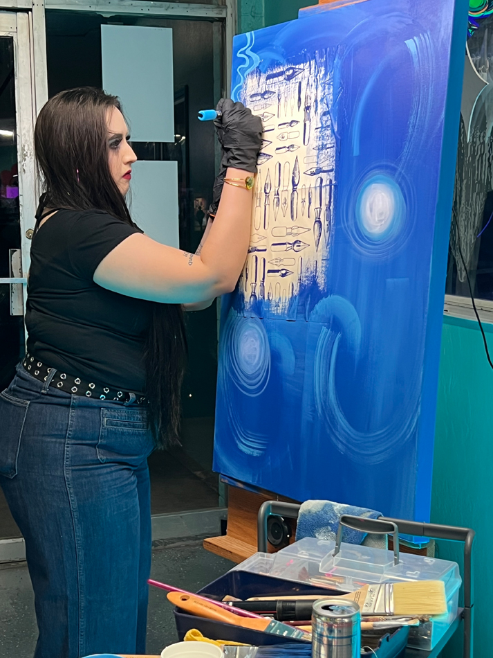 A painter works at a vivid blue canvas with a repeating quill motif. Alternative arts hub Danelle Plaza in Tempe, Arizona, faces uncertainty amid redevelopment plans.