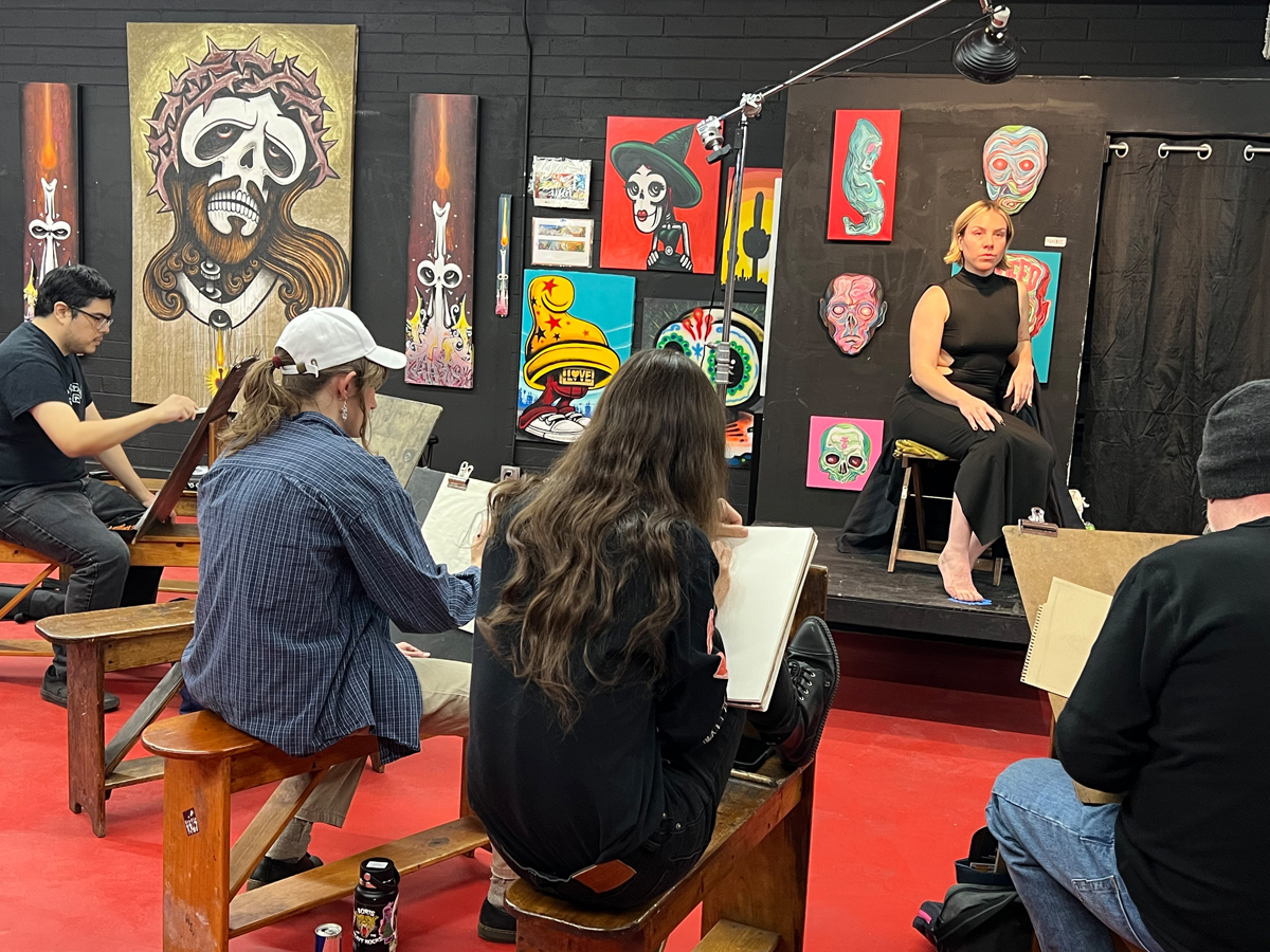 A woman clad in black poses onstage for a life drawing session with numerous artists. Colorful figurative art fills the wall behind her.