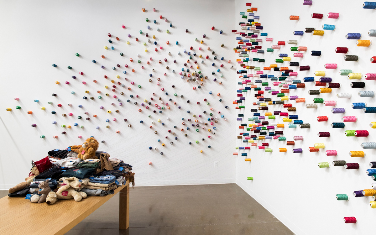 Countless wall-mounted spools of thread connect to a pile of textiles on a bench in an art installation by Lee Mingwei.