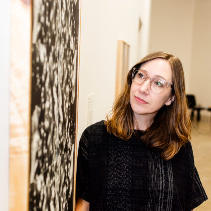 A bespectacled woman dressed in black closely examines a framed wall artwork. A profile of Kimball Art Center curator Nancy Stoaks.