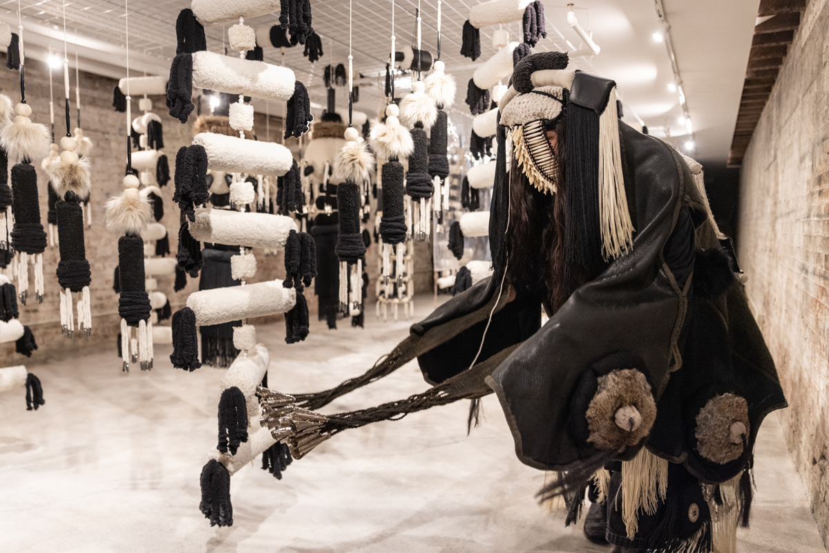 Eric-Paul Riege (Diné/Navajo), decked out in sculptural regalia, performs next to a suspended fiber installation in his first New York solo show.