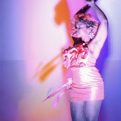 A woman dressed in a frilly pink dress dances against a pink-and-purple backdrop. Artwork by India Sky Davis from the Broken Boxes exhibition.