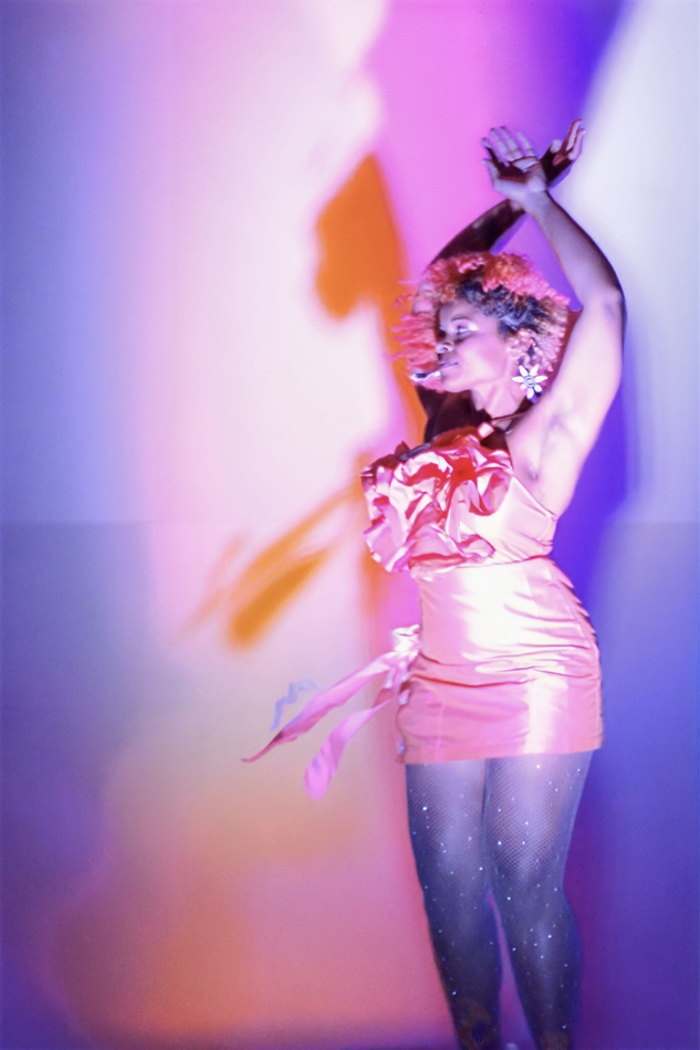 A woman dressed in a frilly pink dress dances against a pink-and-purple backdrop. Artwork by India Sky Davis from the Broken Boxes exhibition.