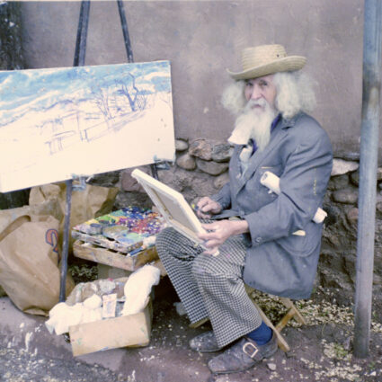 A man sits at an easel, with an adobe wall in the background. Tommy Macaione was a prominent Santa Fe artist.