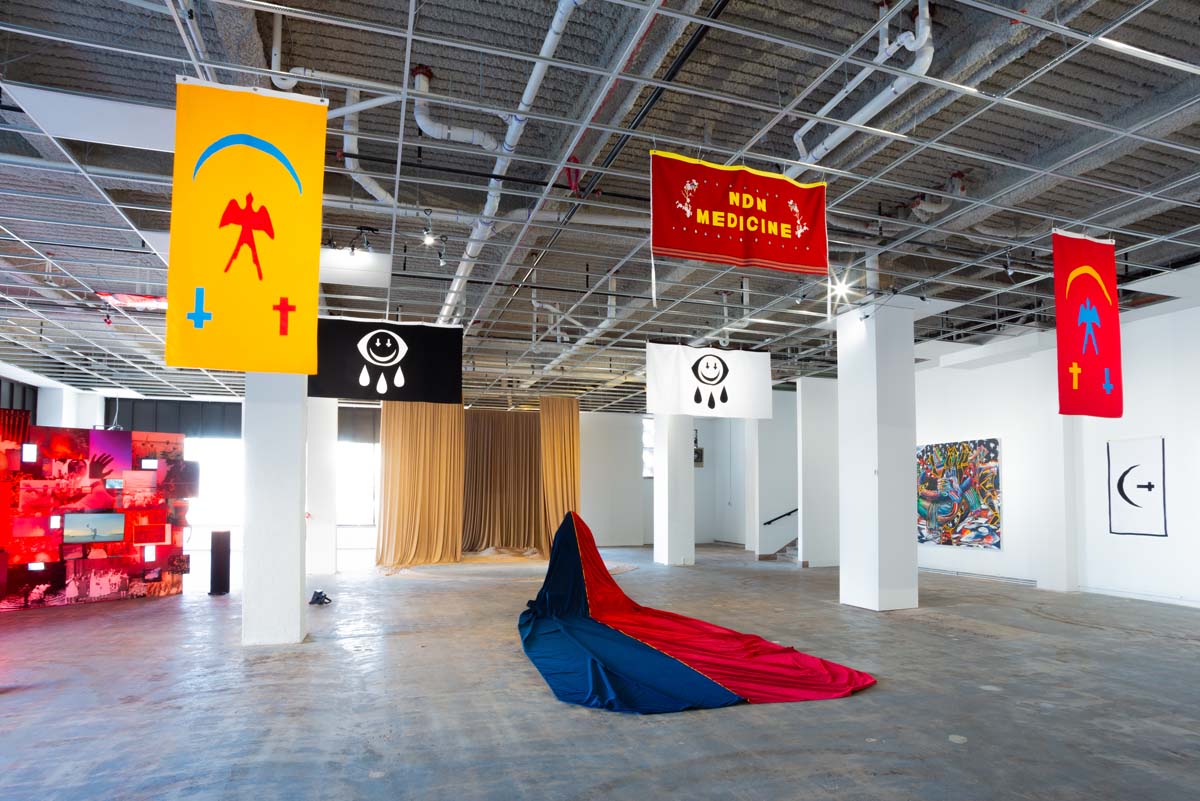 Installation view of Drift///Hold in Tulsa, with artist flags mounted on the ceiling and a suspended, long train of fabric