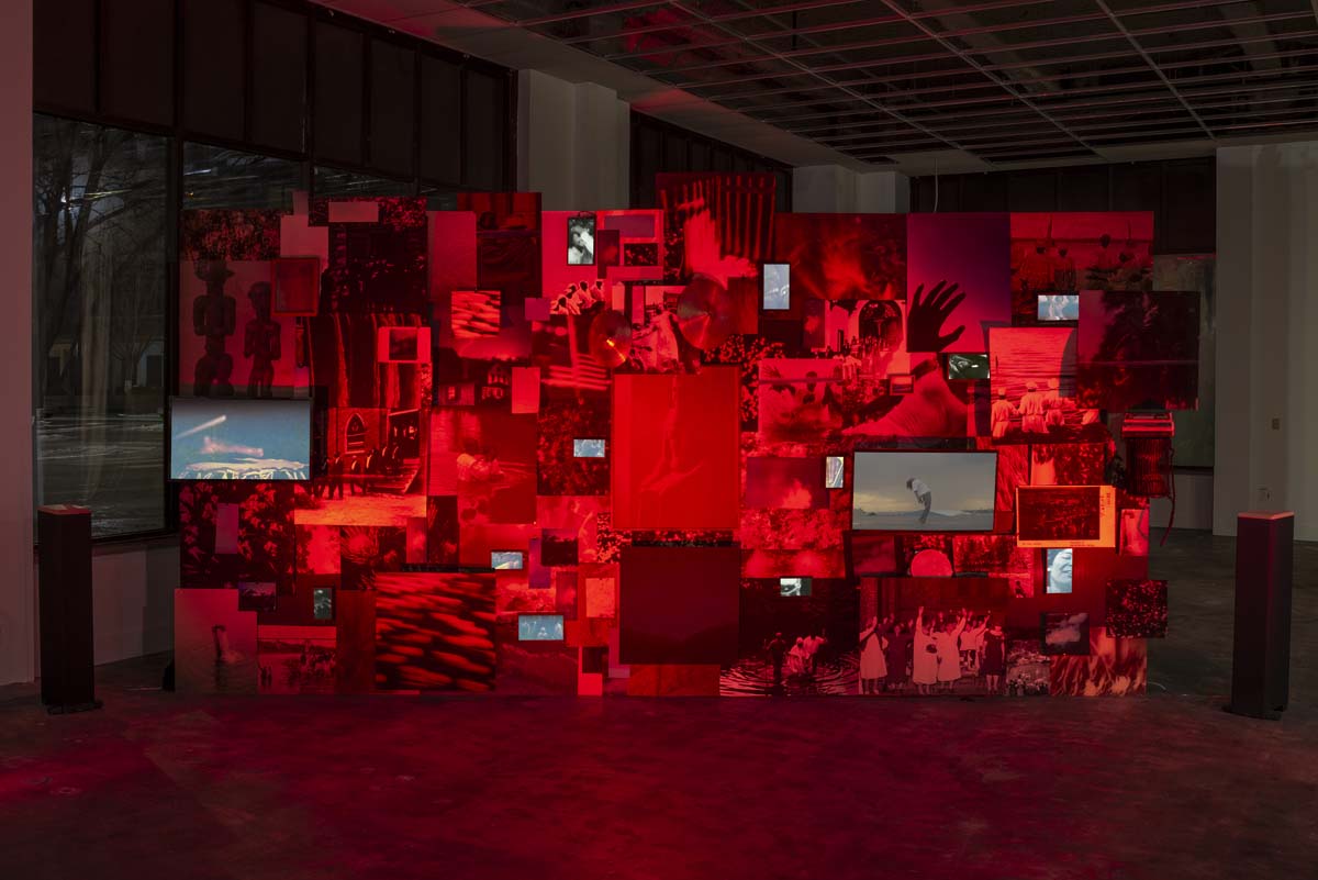 A billboard-sized standing collage bathed in red light, composed of variously-sized prints and video screens by Le'Andra LeSeur