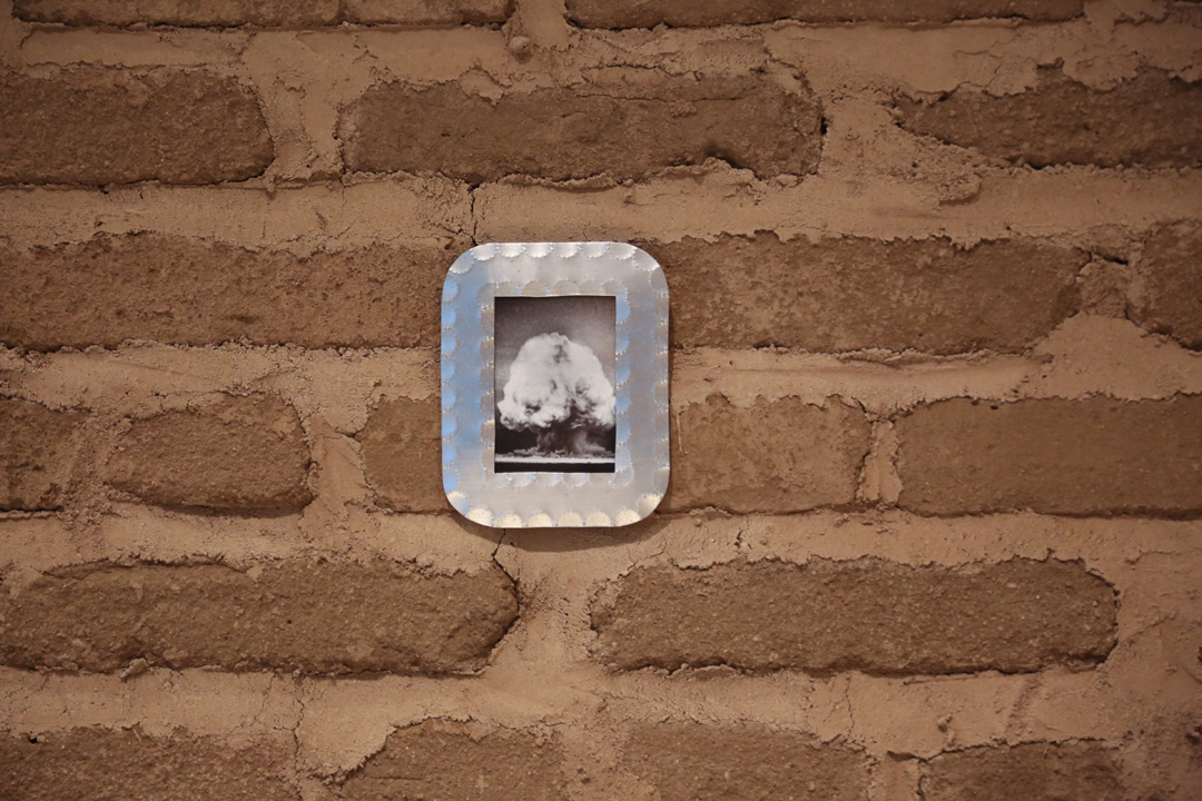 Detail of an installation by Joanna Keane Lopez featuring a brick wall with a photo of a mushroom cloud framed in tinwork. The New Mexico and California-based artist will participate in the SITE Santa Fe International, Once Within a Time.