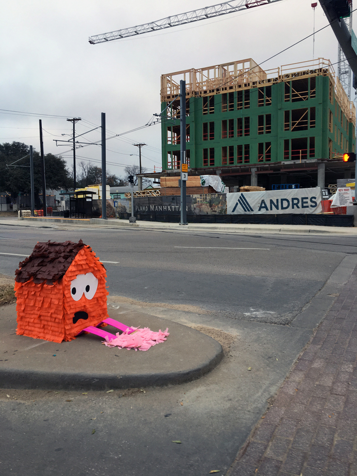 A house-shaped pinatas sits on a Dallas street corner looking distressed. A new development project rises behind it. Art projects from the front lines of the Texas Housing Crisis.