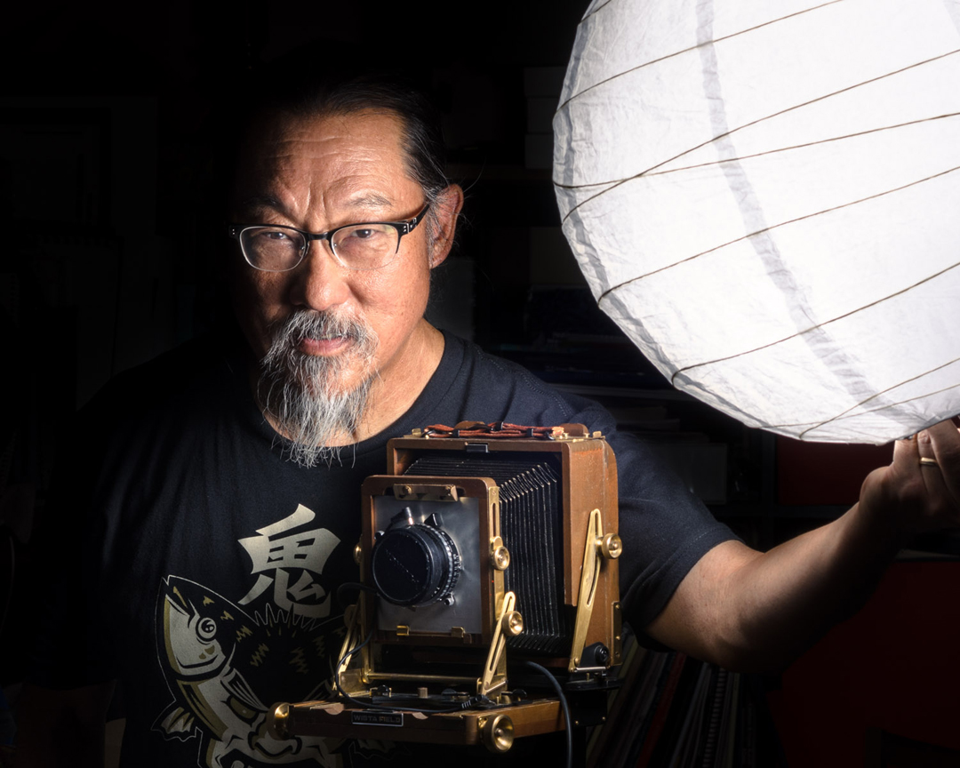Artist Dean Terasaki stands behind a vintage camera holding a globe light.