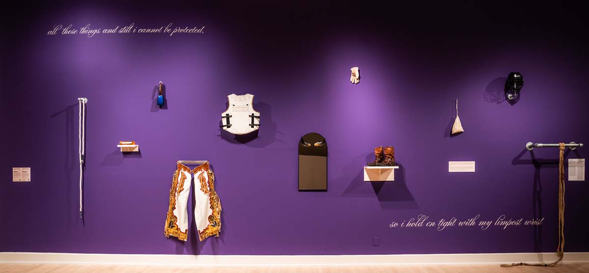 Installation view of a purple wall with a leather vest reading “Queer Rider” on the back in jeweled letters; chaps adorned with gold, red, and blue fringe; rope; glycerin soap; worn gloves; and other items