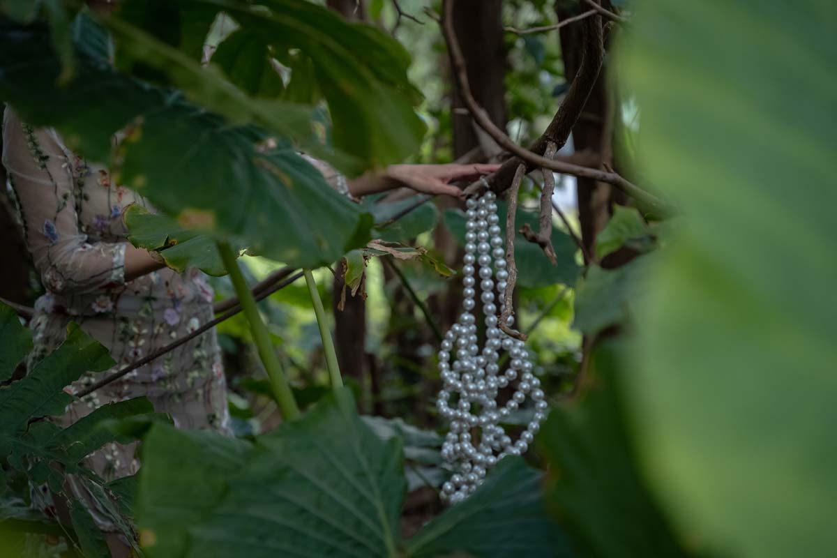 Video still from Lu Wei's Mirrors, showing a hand emerging from dense jungle foliage, holding a large strand of pearls.