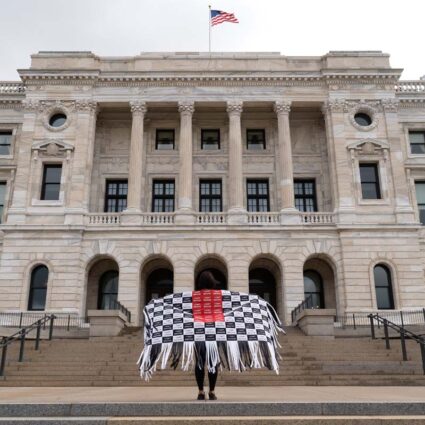 Artist Maggie Thompson standing in front of the Capitol wearing a shawl made of masks with the words I Can't Breathe.