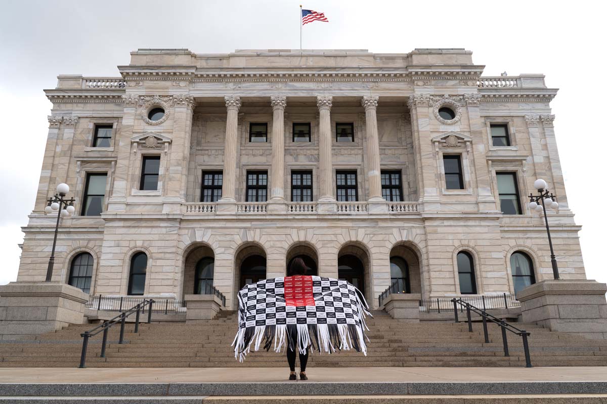 Artist Maggie Thompson standing in front of the Capitol wearing a shawl made of masks with the words I Can't Breathe.