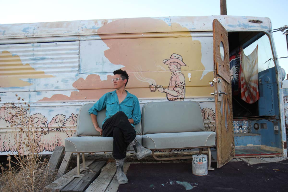 Home of the Brave founder Eileen Muza, sitting on a bench outside of a bus with a mural painted on the outside.