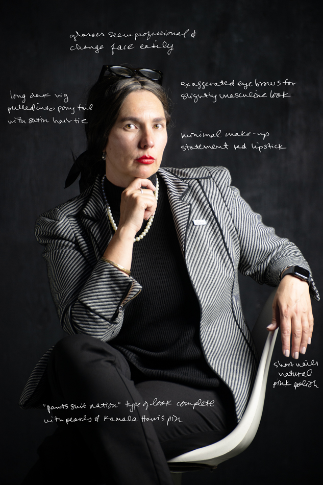 Photograph of artist jesse lovell in one of their disguises named Chelsea, featuring a woman in a brunette wig wearing a grey blazer, sitting in a chair with their hand under their chin.
