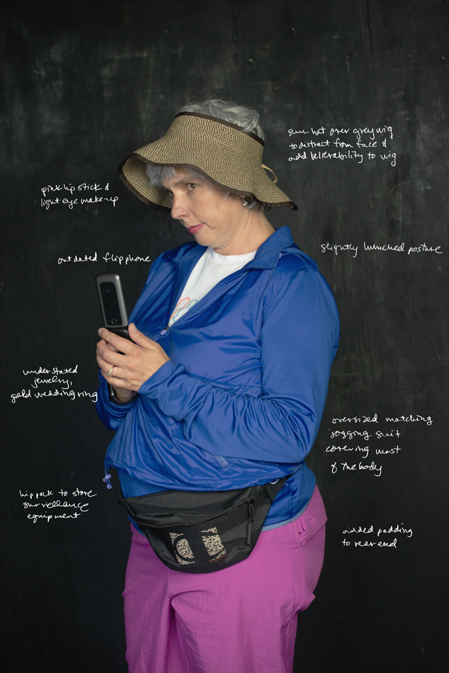 Photograph of artist jesse lovell in one of their disguises named "Theresa" as a "nosy neighbor" type figure. Artist wears a grey wig, wide-brimmed hat, blue jacket, pink pants, fanny pack, and holds up a cell phone in a position that looks like it's recording a video.