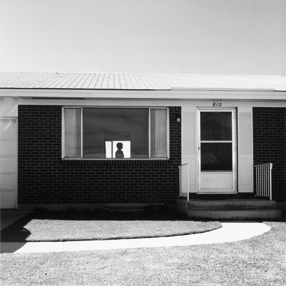 Black and white photo by Robert Adams of a home in Colorado Springs, with the silhouette of a woman visible through the window.