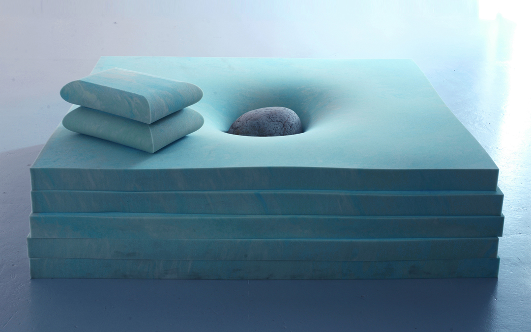 Sculpture of a stone sinking into green memory foam.