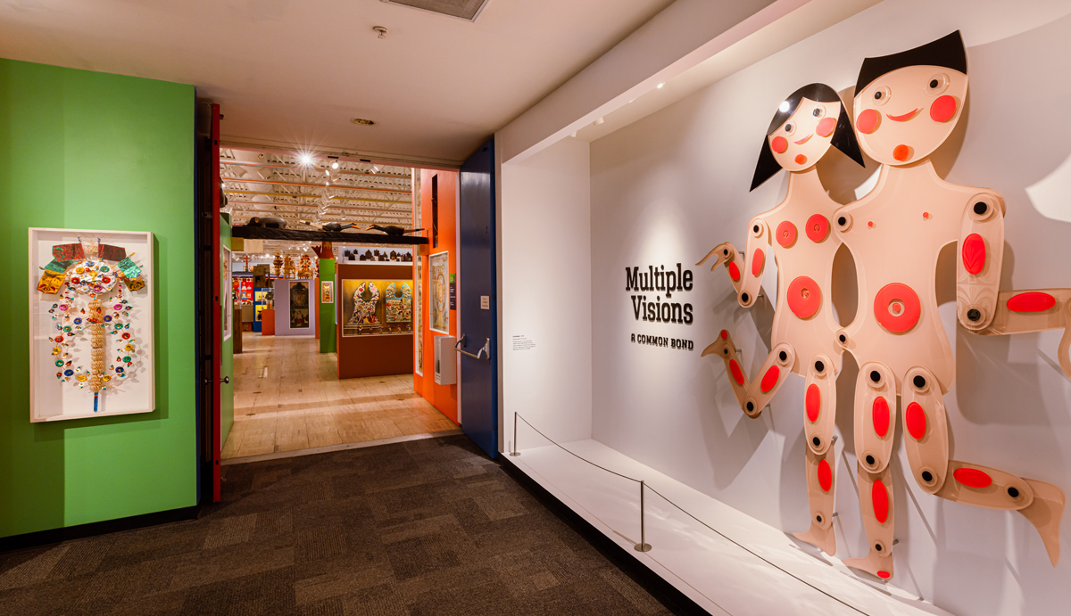 The title wall of Multiple Visions: A Common Bond, showing two dancing doll figures with jointed limbs. The exhibition of folk art was designed by Alexander Girard.