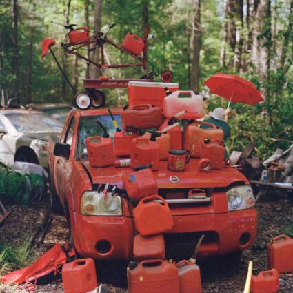 Photograph of an assemblage of red items, mostly gas cans, on a red junk car.
