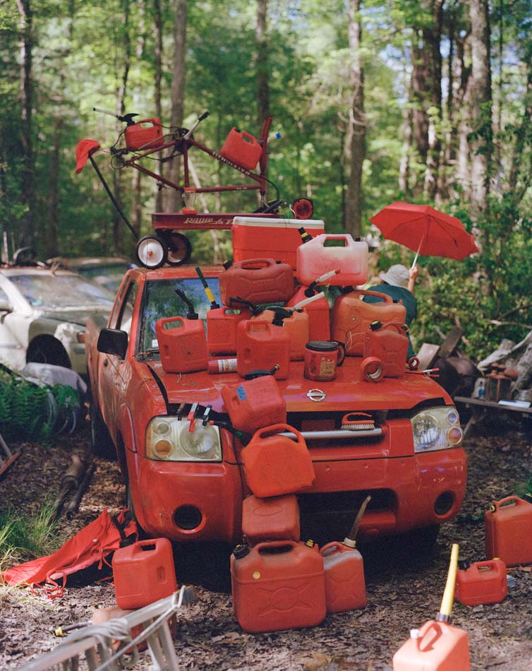 Photograph of an assemblage of red items, mostly gas cans, on a red junk car.