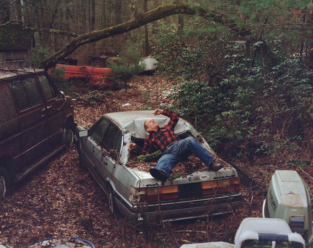 Photograph of a man reclining on top of a junk car in an overgrown wood.