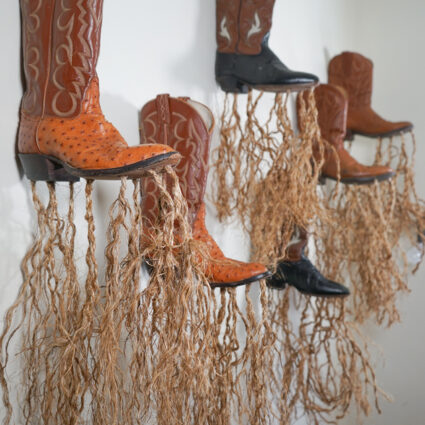 Photo of six brown cowboy boots mounted on a white mall, with strands of loosely braided rope hanging from each boot. Installation by artist José Villalobos at Tucson Museum of Art.