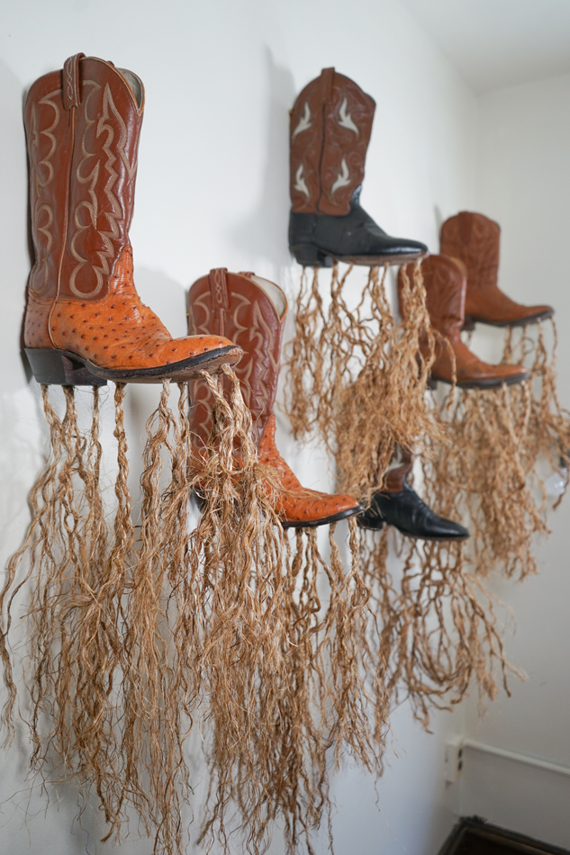 Photo of six brown cowboy boots mounted on a white mall, with strands of loosely braided rope hanging from each boot. Installation by artist José Villalobos at Tucson Museum of Art.