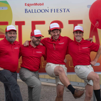 Men dressed as corporate spokespeople do a can-can kick in front of an oversized license plate that reads "ExxonMobil Balloon Fiesta."