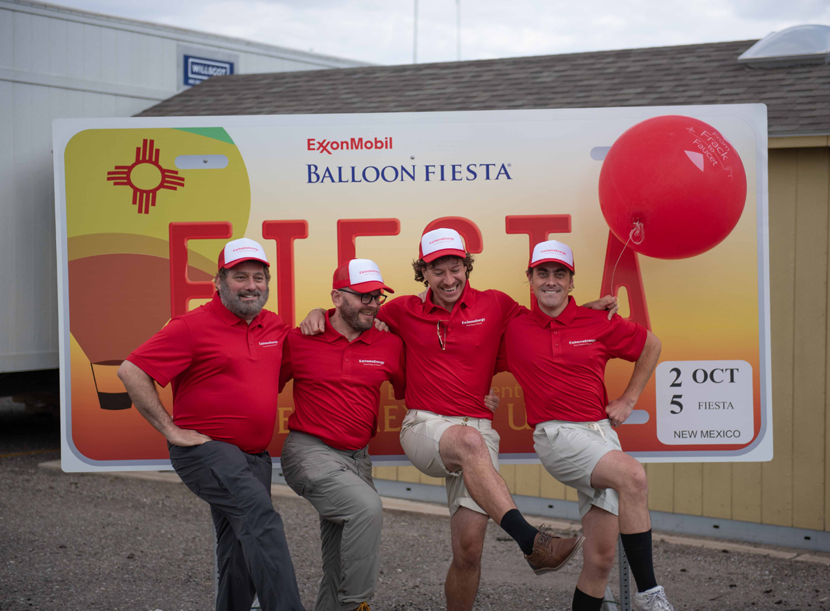 Men dressed as corporate spokespeople do a can-can kick in front of an oversized license plate that reads 