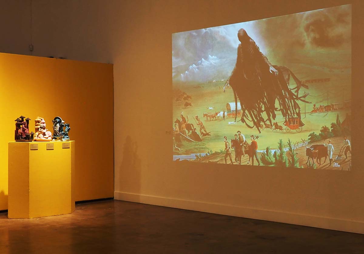 Installation view with Klee Benally's digital gif of the Dark Mark of Manifest Destiny projected on the wall, with ceramic figures by Jeff Schmuki on yellow pedestals.