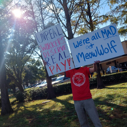 A Meow Wolf Grapevine worker participating in a labor strike holds up two posters. In his right hand, the poster reads "We're all MAD at Meow Wolf". In his left hand, the poster reads "UNREAL WORKERS DESERVE REAL PAY!"