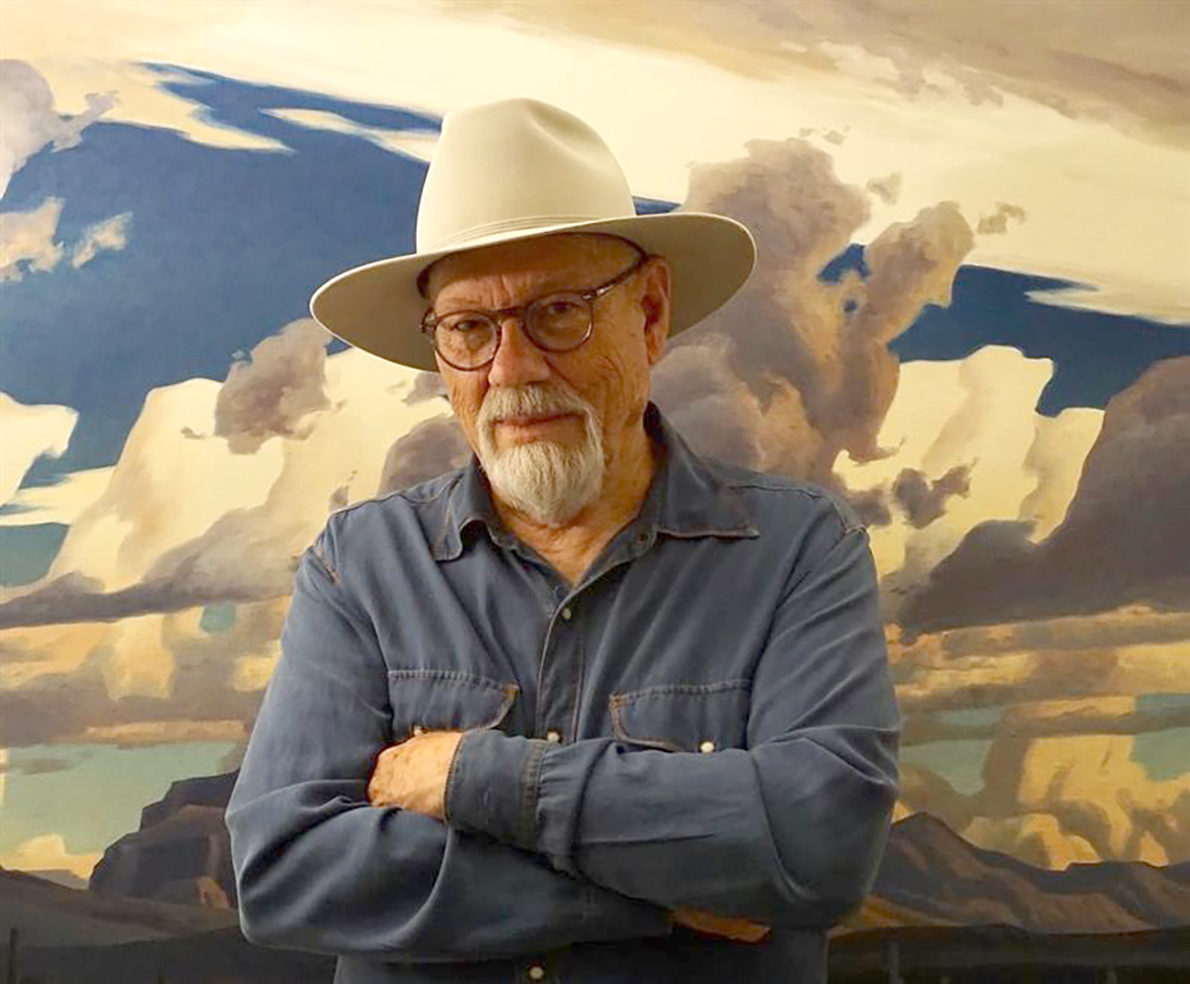 Portrait of late Arizona artist Ed Mell in 2020. He's wearing a cowboy hat and one of his iconic landscape paintings is in the background.