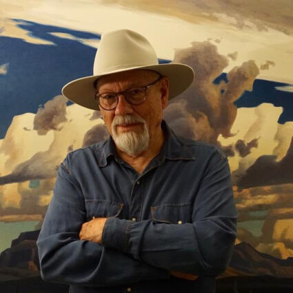 Portrait of late Arizona artist Ed Mell in 2020. He's wearing a cowboy hat and one of his iconic landscape paintings is in the background.