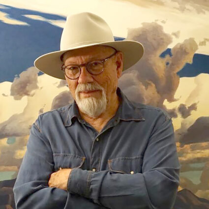 Portrait of late Arizona artist Ed Mell in 2020. He's wearing a cowboy hat and one of his iconic landscape paintings is in the background.